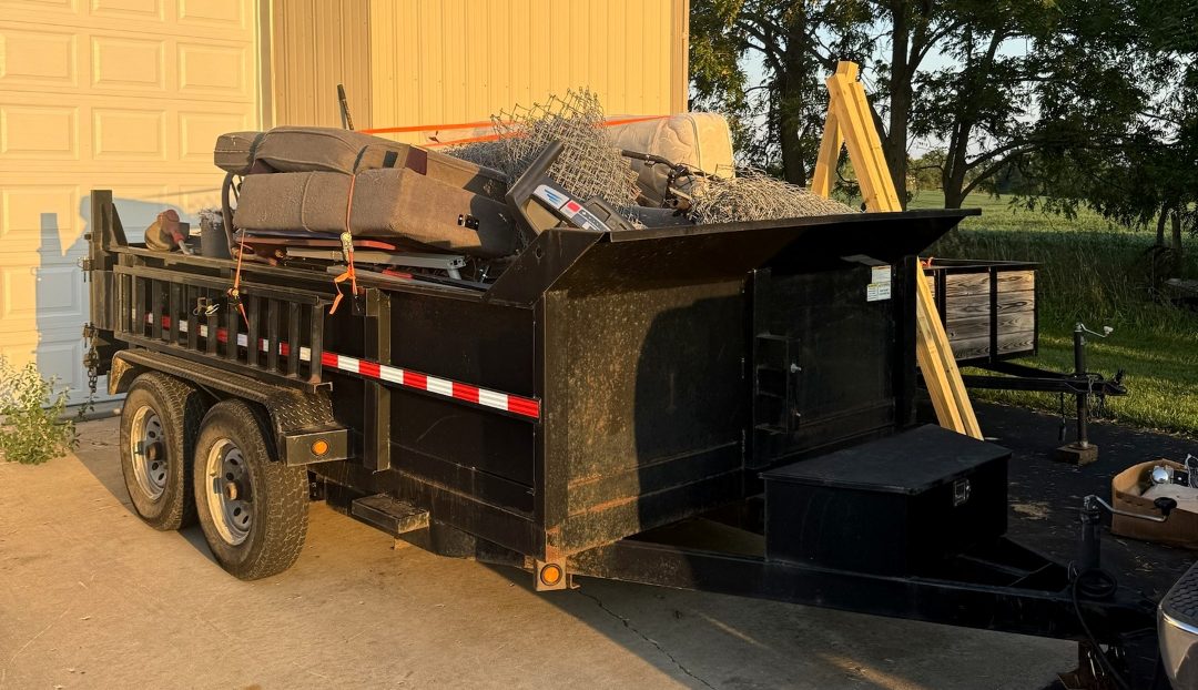 A black tandem-axle dump trailer piled with old furniture, cushions and chain-link fencing